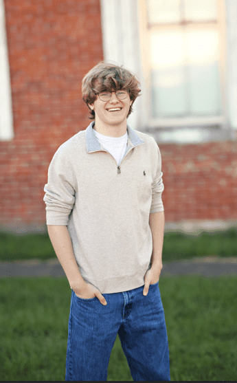Smiling young man in a beige sweater standing outdoors near a brick wall.